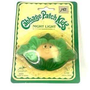 May include: A Cabbage Patch Kids night light featuring a smiling Cabbage Patch Kid face with green cabbage leaves. The packaging is yellow with green trim and says "Cabbage Patch Kids Night Light" and "1984 Original Appalachian Artworks, Inc."