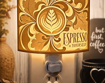 Könnte beinhalten: Ein Stecker-Nachtlicht mit einem warm beleuchteten Schirm, der ein lasergeschnittenes Design einer Kaffeetasse mit Latte-Art, geschwungenen Blattmustern und dem Text Espresso + Yourself zeigt, montiert auf einer Standard-Steckdose.