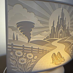 May include: A curved, illuminated white lamp featuring a relief carving of a landscape with a tornado, a castle in the distance, a winding path, and a pair of high-heeled shoes in the foreground, all set against a sunburst background.