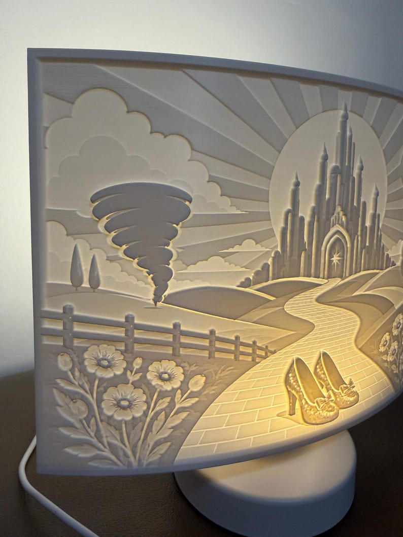 May include: A curved, illuminated white lamp featuring a relief carving of a landscape with a tornado, a castle in the distance, a winding path, and a pair of high-heeled shoes in the foreground, all set against a sunburst background.