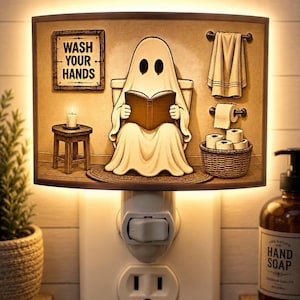 May include: A rectangular plug-in night light featuring a cartoon ghost sitting on a toilet reading a book. The scene includes a sign that reads WASH YOUR HANDS, a towel, a roll of toilet paper, a basket of toilet paper rolls, and a small stool with a candle.