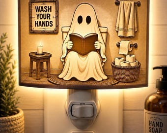 Könnte beinhalten: Ein rechteckiges Nachtlicht für die Steckdose mit einem Cartoon-Geist, der auf einer Toilette sitzt und ein Buch liest. Das Bild zeigt ein Schild mit der Aufschrift WASH YOUR HANDS, ein Handtuch, Toilettenpapier, einen Korb mit Rollen und einen Hocker.