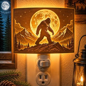 May include: A rectangular night light featuring a warm, glowing silhouette of Bigfoot walking across a mountain ridge in front of a large, bright full moon. The scene includes detailed pine trees and jagged mountain peaks, all illuminated from behind.