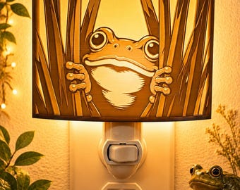 Könnte beinhalten: Ein Steckdosen-Nachtlicht mit einem rechteckigen Schirm, der einen ausgeschnittenen Frosch zeigt, der durch hohes Gras schaut. Die Lampe strahlt ein warmes, goldenes Licht hinter der Illustration aus und betont die großen Augen des Frosches.