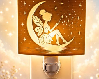 Könnte beinhalten: Ein rechteckiges Nachtlicht für die Steckdose mit warmem goldenem Licht. Die Frontplatte zeigt die Silhouette einer geflügelten Fee, die auf einer Mondsichel sitzt und einen Zauberstab hält, der einen Sternenschweif aussendet. Das Licht steckt in einer Steckdose.