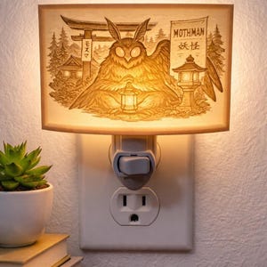 May include: A night light featuring a detailed illustration of the Mothman with Japanese text and imagery. The lamp is plugged into a wall outlet and emits a warm, soft glow. A small succulent in a white pot sits nearby.