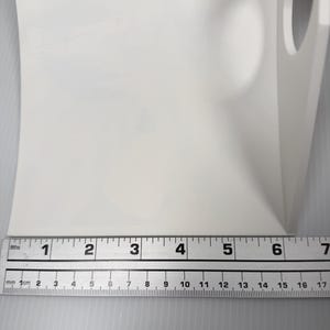 May include: A white L-shaped plastic bracket with a circular cutout on the vertical side. A metal ruler is positioned along the base, showing a width of approximately 6.5 inches. The object has a smooth, matte finish and a rigid, angular structure.