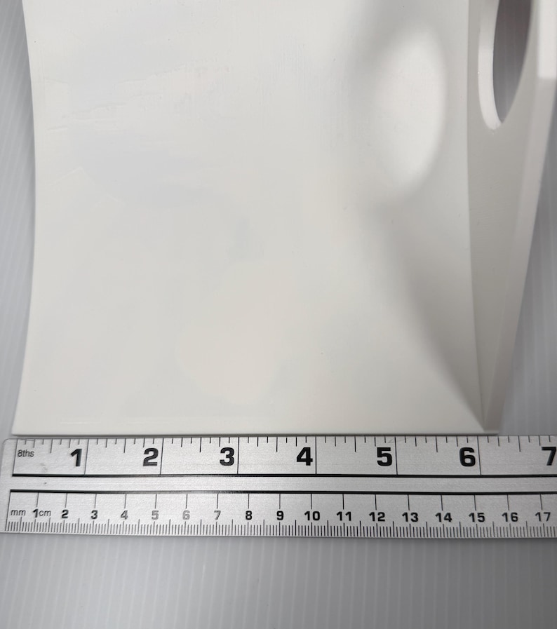 May include: A white L-shaped plastic bracket with a circular cutout on the vertical side. A metal ruler is positioned along the base, showing a width of approximately 6.5 inches. The object has a smooth, matte finish and a rigid, angular structure.