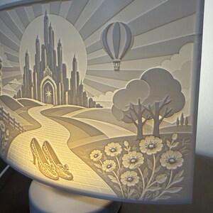 May include: A curved, illuminated white lamp featuring a relief carving of a castle, a hot air balloon, a brick path, a pair of high-heeled shoes, and flowers. The scene is backlit, creating a warm glow that highlights the intricate details.