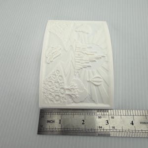 Pode incluir: Um molde de silicone branco retangular com um design em relevo de um castelo, um bal&atilde;o de ar quente e &aacute;rvores. Uma r&eacute;gua de metal est&aacute; posicionada na parte inferior, mostrando que o molde tem cerca de 8,9 cm de largura na base.
