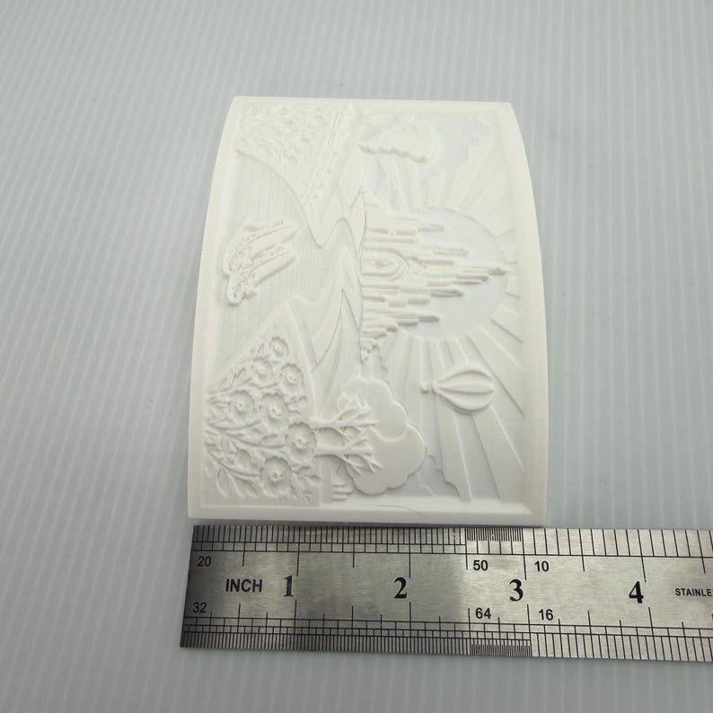 Pode incluir: Um molde de silicone branco retangular com um design em relevo de um castelo, um bal&atilde;o de ar quente e &aacute;rvores. Uma r&eacute;gua de metal est&aacute; posicionada na parte inferior, mostrando que o molde tem cerca de 8,9 cm de largura na base.