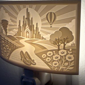 Pode incluir: Uma luz noturna retangular de tomada com uma cena esculpida em relevo de um castelo, um bal&atilde;o de ar quente, um caminho de tijolos e um par de sapatos em primeiro plano. A superf&iacute;cie esbranqui&ccedil;ada &eacute; iluminada por tr&aacute;s com um brilho suave.