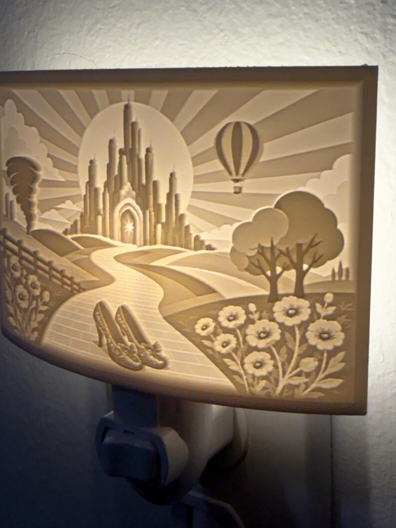 Pode incluir: Uma luz noturna retangular de tomada com uma cena esculpida em relevo de um castelo, um bal&atilde;o de ar quente, um caminho de tijolos e um par de sapatos em primeiro plano. A superf&iacute;cie esbranqui&ccedil;ada &eacute; iluminada por tr&aacute;s com um brilho suave.