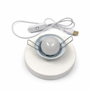 May include: A white circular base featuring a central recessed metal housing with a round white light bulb. Two silver metal spring clips extend from the sides of the housing. A white power cord with a USB connector and an inline push-button switch.