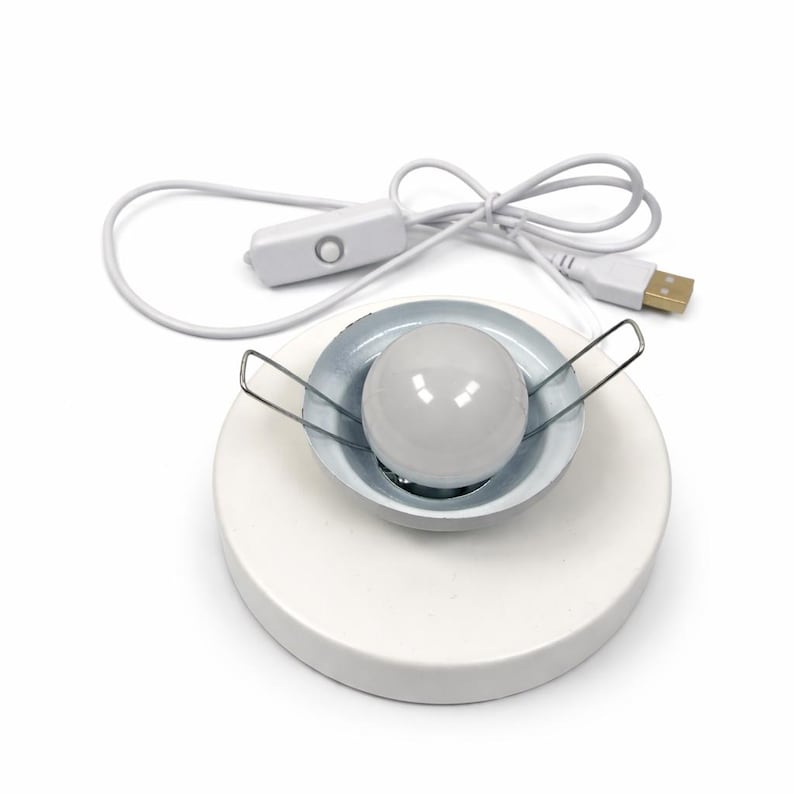 May include: A white circular base featuring a central recessed metal housing with a round white light bulb. Two silver metal spring clips extend from the sides of the housing. A white power cord with a USB connector and an inline push-button switch.