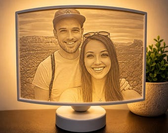 Könnte beinhalten: Eine rechteckige, beleuchtete 3D-gedruckte Lithophanie-Lampe mit dem Porträt zweier lächelnder Personen. Die Lampe hat einen weißen Rahmen und einen runden Sockel und strahlt ein warmes, goldenes Licht aus, das die Gesichtszüge hervorhebt.