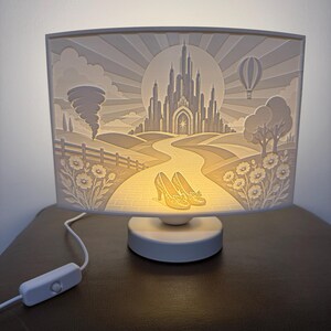 May include: A rectangular white lamp featuring a backlit 3D relief scene of a castle at the end of a path, with a pair of yellow shoes in the foreground, a tornado on the left, a hot air balloon on the right, and flowers along the path.