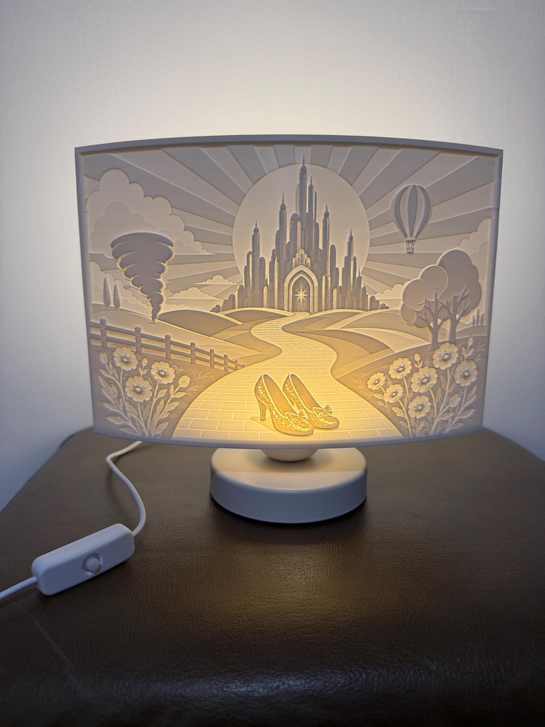 May include: A rectangular white lamp featuring a backlit 3D relief scene of a castle at the end of a path, with a pair of yellow shoes in the foreground, a tornado on the left, a hot air balloon on the right, and flowers along the path.