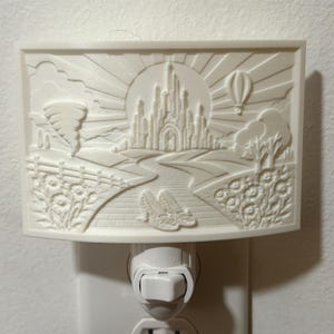 Pode incluir: Uma luz noturna retangular branca com um relevo de um castelo, um tornado, um bal&atilde;o de ar quente e um par de sapatos num caminho de tijolos. A cena inclui raios de sol, nuvens e elementos florais montados numa tomada de parede.