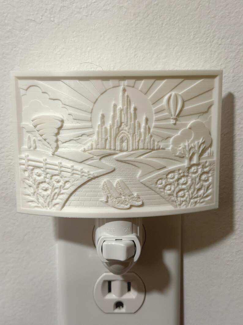 Pode incluir: Uma luz noturna retangular branca com um relevo de um castelo, um tornado, um bal&atilde;o de ar quente e um par de sapatos num caminho de tijolos. A cena inclui raios de sol, nuvens e elementos florais montados numa tomada de parede.