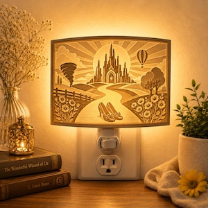 May include: A rectangular plug-in night light featuring a layered paper-cut style illustration of a castle, a tornado, a hot air balloon, and a pair of glittery shoes on a path, all illuminated with a warm glow from behind the scene.