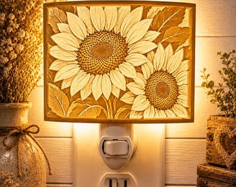 Könnte beinhalten: Ein rechteckiges Steckdosen-Nachtlicht mit einem geschnitzten Sonnenblumenmotiv, bestehend aus zwei großen Blüten und Blättern. Das Licht strahlt warm hinter der texturierten, cremefarbenen Platte hervor, die in einer weißen Wandsteckdose steckt.