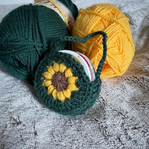 Puede incluir: Un soporte de hilo de ganchillo hecho a mano con un diseño de girasol, en tonos verdes, amarillos y marrones. El soporte está junto a dos ovillos de lana, uno amarillo y otro verde oscuro. El conjunto está sobre una superficie gris texturizada.