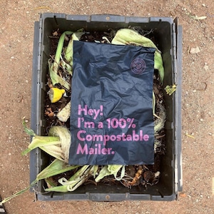 May include: A black compostable mailer bag with the text "Hey! I'm a 100% Compostable Mailer." printed in pink. The bag is sitting in a black compost bin with various organic materials.
