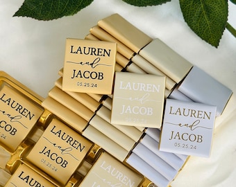 May include: A selection of personalised chocolate bars in gold, ivory, and silver packaging. Each bar is printed with the names "LAUREN and JACOB" and the date "05.25.24". The chocolates are arranged in a gold tray, ideal for wedding favours.
