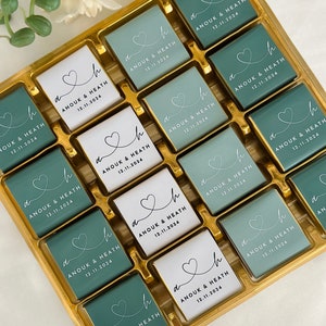 May include: A gold box filled with square chocolates. Some chocolates are teal, others are white. Each chocolate has a heart design and the text "ANOUK & HEATH 12.11.2024" in black. The chocolates are arranged in neat rows.