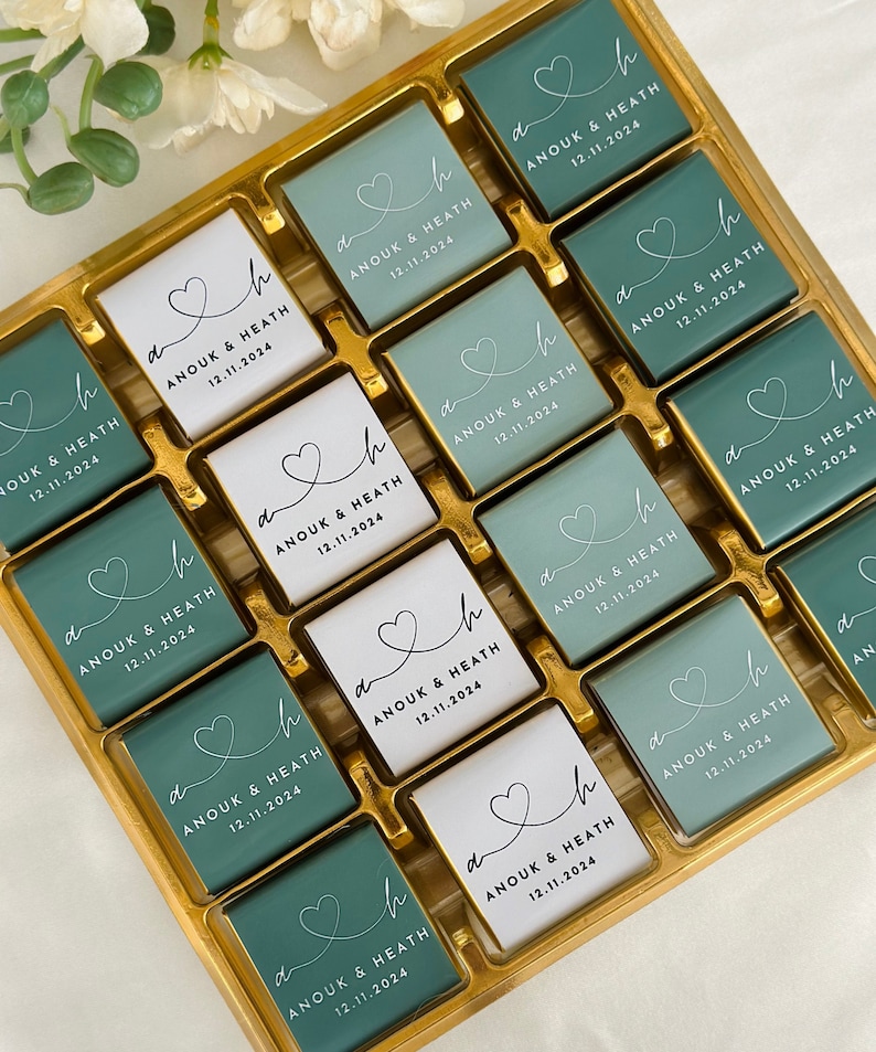 May include: A gold box filled with square chocolates. Some chocolates are teal, others are white. Each chocolate has a heart design and the text "ANOUK & HEATH 12.11.2024" in black. The chocolates are arranged in neat rows.