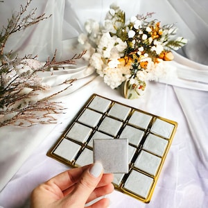 May include: A gold tray with 16 square compartments filled with white, textured, square tiles. One tile is being held in a hand, showing the texture of the tile.