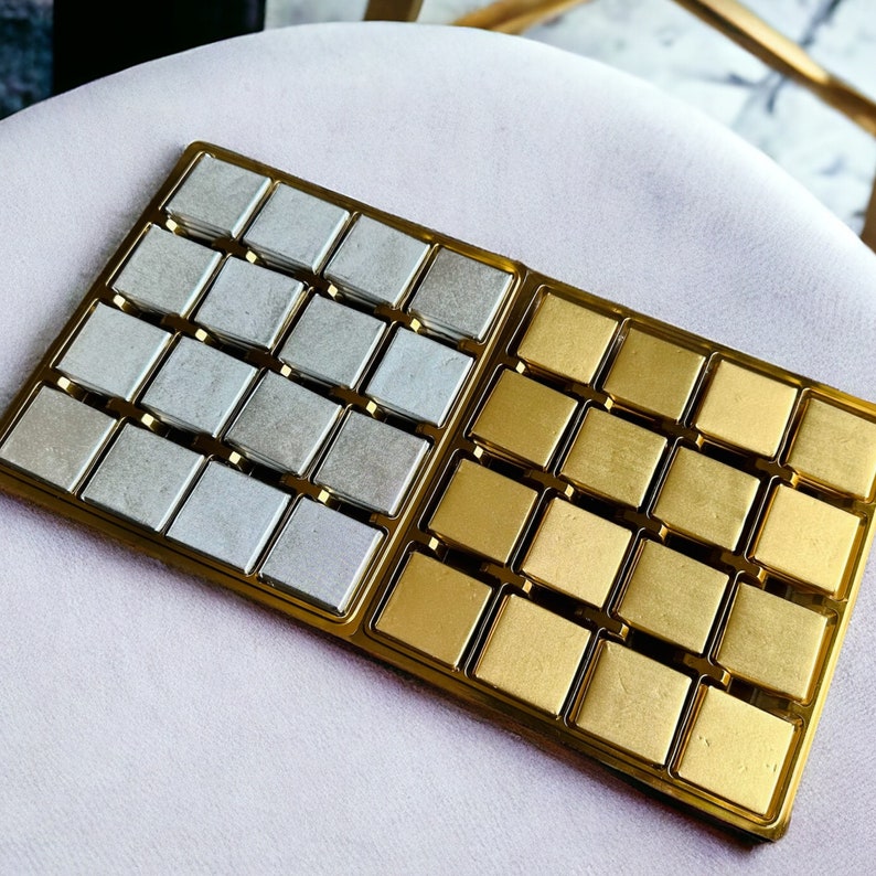 May include: A gold and silver tray with 24 square compartments filled with chocolate squares. The chocolate squares are arranged in two rows of 12. The squares on the left side of the tray are silver, and the squares on the right side of the tray are gold.