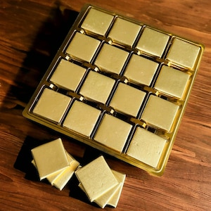 May include: A gold-colored tray with 16 square compartments, each filled with a small, square, gold-colored chocolate. Three additional chocolates are stacked on a wooden surface.