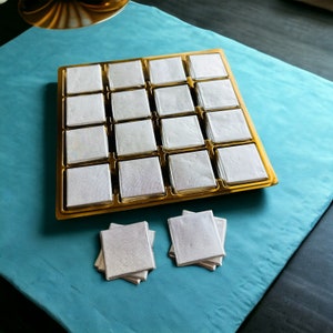 May include: A gold tray with 16 white squares, each with a subtle iridescent sheen. Two stacks of white squares are on a blue surface.
