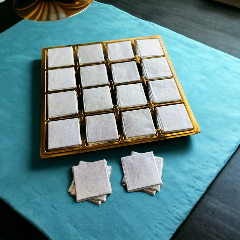 May include: A gold tray with 16 white squares, each with a subtle iridescent sheen. Two stacks of white squares are on a blue surface.