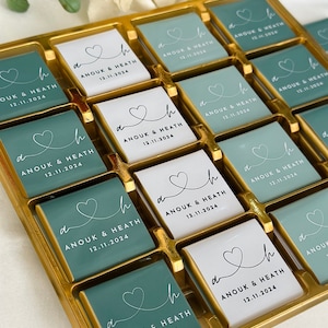 May include: A gold tray filled with square chocolates. Some chocolates are white, others are teal. Each chocolate has a heart design and the text "ANOUK & HEATH 12.11.2024". The chocolates are arranged in rows and columns.