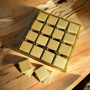 May include: A gold-colored tray with 16 individual compartments, each containing a square-shaped chocolate. The tray is on a wooden surface.