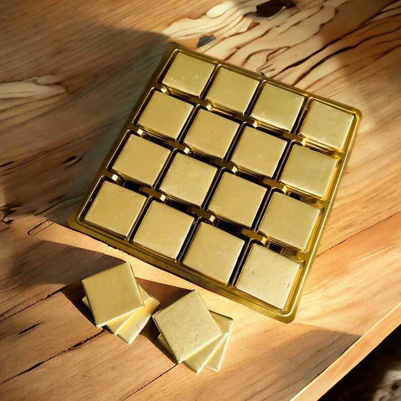 May include: A gold-colored tray with 16 individual compartments, each containing a square-shaped chocolate. The tray is on a wooden surface.
