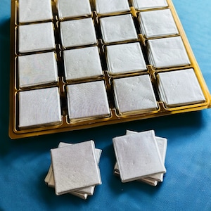 May include: A gold tray with 18 square compartments filled with white shimmery squares. Two stacks of white shimmery squares are on a blue surface.