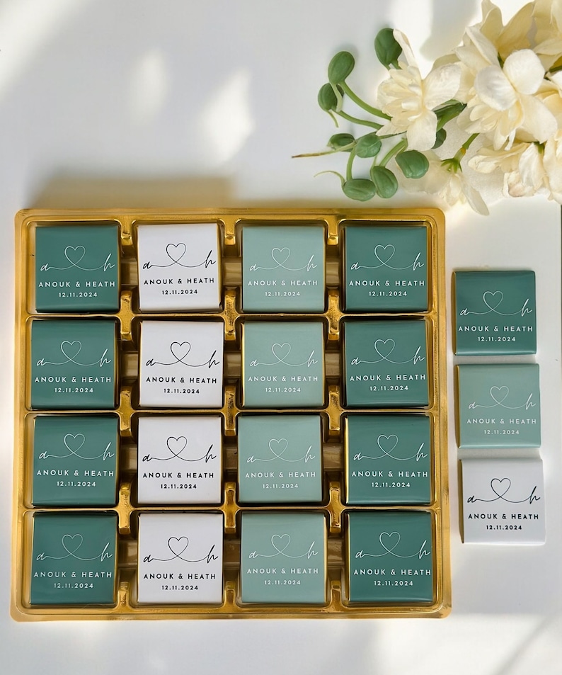 May include: A gold tray filled with square chocolates. Some chocolates are teal, some are white. Each chocolate has a heart design and the text "ANOUK & HEATH 12.11.2024". The chocolates are arranged in rows and columns.