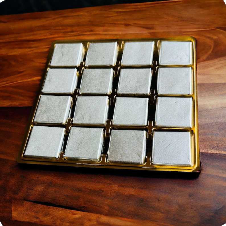 May include: A gold-colored tray with 16 square compartments, each filled with a white, square-shaped item. The tray is on a wooden surface.