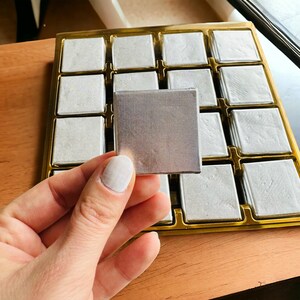 May include: A silver tray with multiple square compartments, each containing a small, square, silver-colored object. The tray is made of gold-colored metal and has a wooden surface underneath.