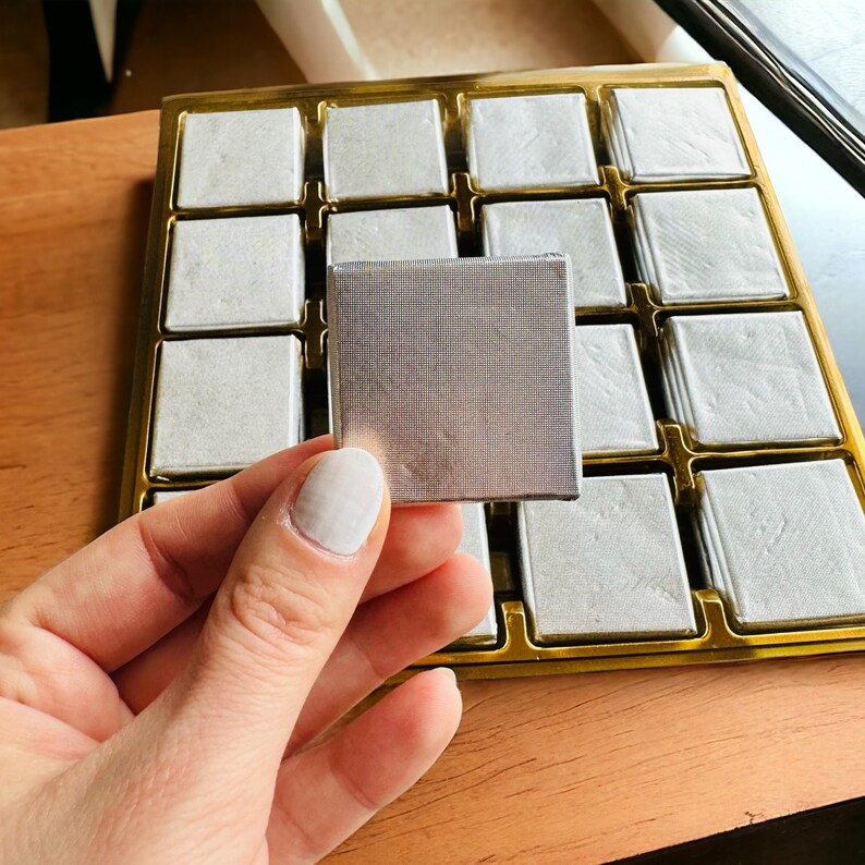 May include: A silver tray with multiple square compartments, each containing a small, square, silver-colored object. The tray is made of gold-colored metal and has a wooden surface underneath.