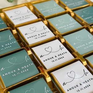 May include: A gold tray filled with square chocolates. Some chocolates are white, others are teal. Each chocolate has a heart design and the text "ANOUK & HEATH 12.11.2024". A wedding favor or gift.