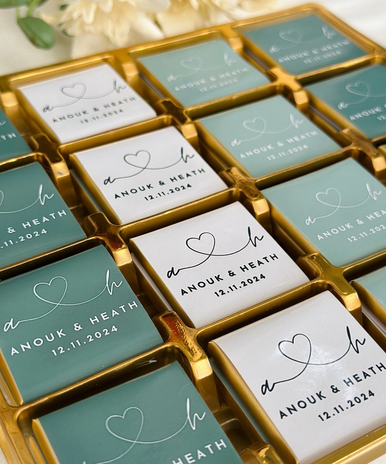 May include: A gold tray filled with square chocolates. Some chocolates are white, others are teal. Each chocolate has a heart design and the text "ANOUK & HEATH 12.11.2024". A wedding favor or gift.