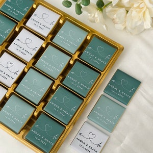 May include: A gold tray filled with square chocolates. The chocolates are teal and white, each with a heart design and the text "ANOUK & HEATH 12.11.2024" in white script. The chocolates are arranged in rows.