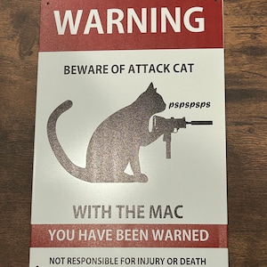 May include: A white metal sign with red lettering that reads "WARNING BEWARE OF ATTACK CAT WITH THE MAC YOU HAVE BEEN WARNED NOT RESPONSIBLE FOR INJURY OR DEATH". The sign features a black silhouette of a cat holding a gun.