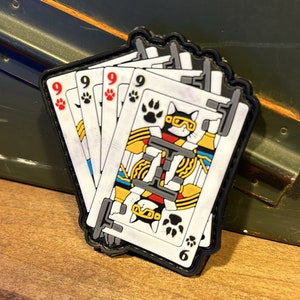 May include: A black and white patch with a cartoon cat wearing goggles and a yellow and red outfit. The cat is holding a gun and is surrounded by playing cards with the number 9 on them. The patch has a black paw print design and the number 6 on the bottom.
