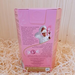 May include: A pink and white box containing a chocolate truffle. The box features an image of a chef holding a whisk and a bowl of chocolate. The text on the box reads "Wann immer, wo immer Sie LINDOR geniessen - es ist ein magischer Moment. Wenn die feine Chocoladenh&uuml;lle bricht, verf&uuml;hrt die unendlich zartschmelzende F&uuml;llung Ihre Sinne und tr&auml;gt Sie sanft davon - ein Moment  puren Chocoladengl&uuml;cks. Es ist Ihr Moment. Mit LINDOR - kreiert von den Lindt Ma&icirc;tres Chocolatiers. Liebe und Leidenschaft f&uuml;r Chocolade seit 1845." The text on the bottom of the box reads "HIGHLY MELTABLE KEEP COOL QUALITY GUARANTEE MANUFACTURED BY LINDT & SPRUNGLI AG LINDT & SPRUNGLI (UK) LTD LINDT SUSTAINABILITY 02 2025 L294400"